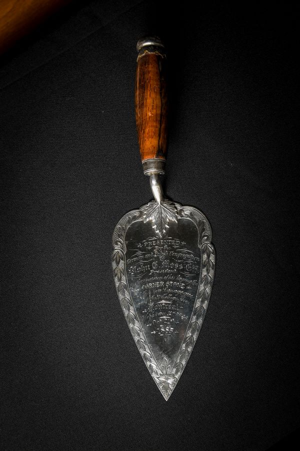 Une truelle en métal gravée sur une surface noire. L’inscription mentionne : « présenté par la Congrégation allemande et polonaise à John E. Moss, président, à l’occasion de la pose de la première pierre de la nouvelle synagogue sur l’avenue McGill College à Montréal. Heshvan, 12e, 5656. 21 octobre 1885 ». 