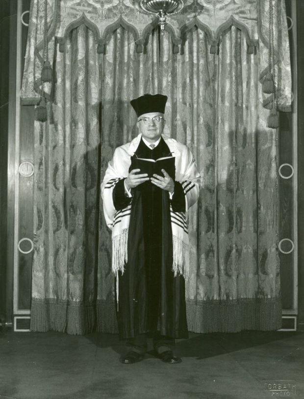 Une photographie en noir et blanc de le chantre Nathan Mendelson debout devant l’arche de la synagogue. Il porte un chapeau noir de chantre, des vêtements cérémoniels noirs, et un talit, tout en tenant un livre de prières.