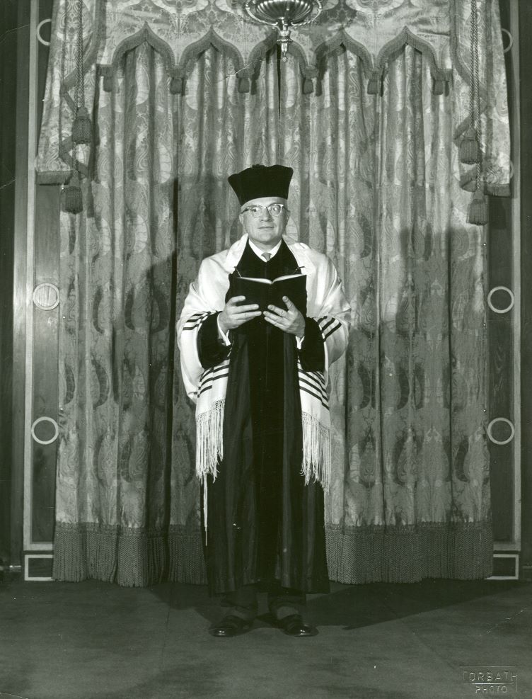 Une photographie en noir et blanc de le chantre Nathan Mendelson debout devant l’arche de la synagogue. Il porte un chapeau noir de chantre, des vêtements cérémoniels noirs, et un talit, tout en tenant un livre de prières.