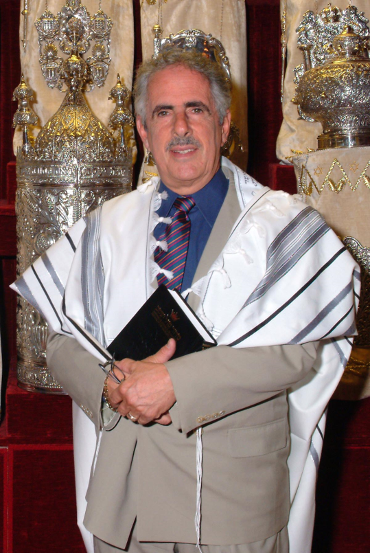 Un homme vêtu d’un châle de prière blanc, debout devant les rouleaux de la Torah dans l’arche de la synagogue.