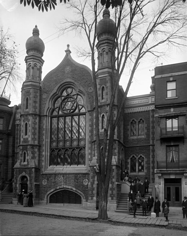 Une photographie en noir et blanc de l’extérieur de la synagogue construite dans le style néo-mauresque. Un groupe d’individus rassemblés dans l’escalier de la synagogue.