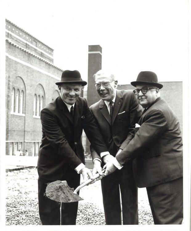 une photographie en noir et blanc de trois hommes en complet qui échangent une pelle.