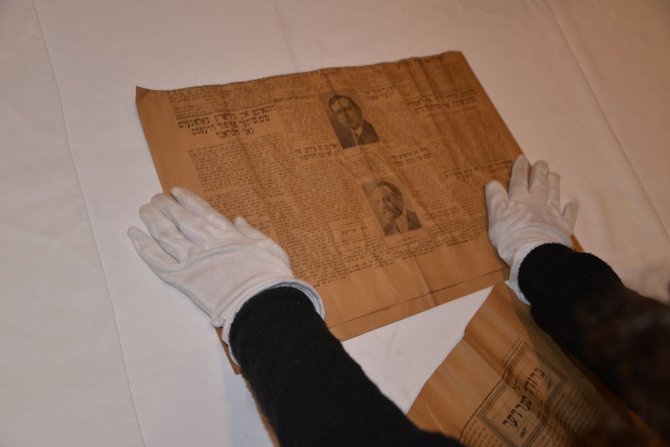 Un ancien journal yiddish déposé sur une table consulté par un individu portant des gants blancs.