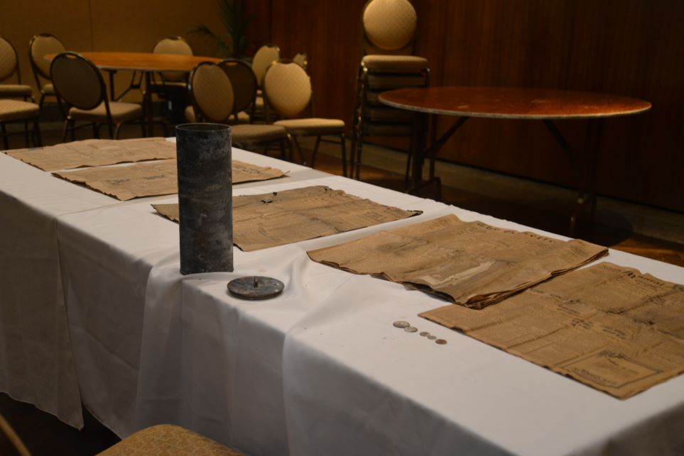 Plusieurs journaux anciens, une capsule temporelle dans un cylindre en laiton et cinq pièces de monnaie présentées sur une table avec une serviette blanche.