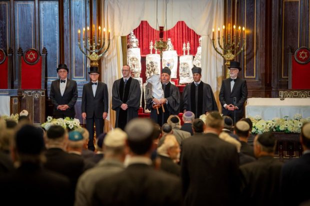 Un groupe d’hommes vêtus de tenues formelles et cérémonielles se tient devant la bimah (tribune) de la synagogue pour la cérémonie de réouverture.