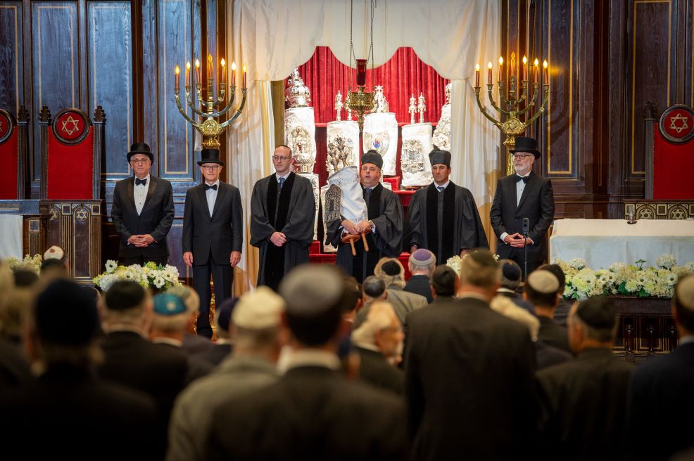Un groupe d’hommes vêtus de tenues formelles et cérémonielles se tient devant la bimah (tribune) de la synagogue pour la cérémonie de réouverture.