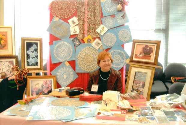 Une femme souriante est assise derrière une table. Derrière elle, des centres de tables crochetés, des cartes de souhaits et des toiles sont exposés. Devant elle, des articles crochetés et des cartes sont déposés sur une table.