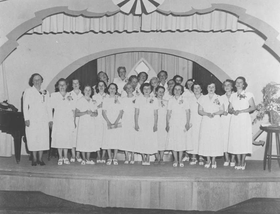 Photographie en noir et blanc d’une vingtaine de femmes vêtues de blanc et portant des boutonnières de fleurs qui sont regroupées sur une scène. On aperçoit un piano à leur gauche et un bouquet de fleurs sur un tabouret à leur droite.