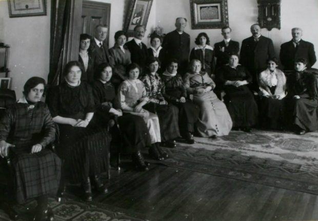 Quatorze femmes vêtues de jupes ou de robes longues et six hommes vêtus de costumes sont installés dans le coin d’une pièce. Les femmes de la rangée du devant sont toutes assises. Les hommes et les femmes de la deuxième rangée sont debout.