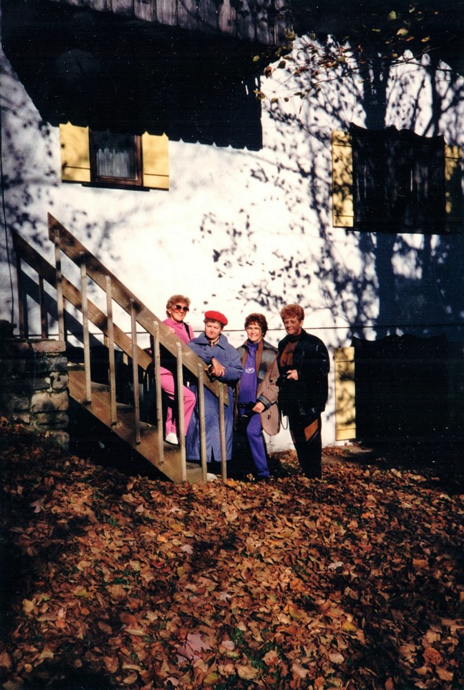 Quatre femmes sont regroupées à l’extérieur, au pied d’un escalier devant un bâtiment blanc dont les fenêtres ont des volets jaunes. Devant elles se trouve un tapis de feuilles mortes.