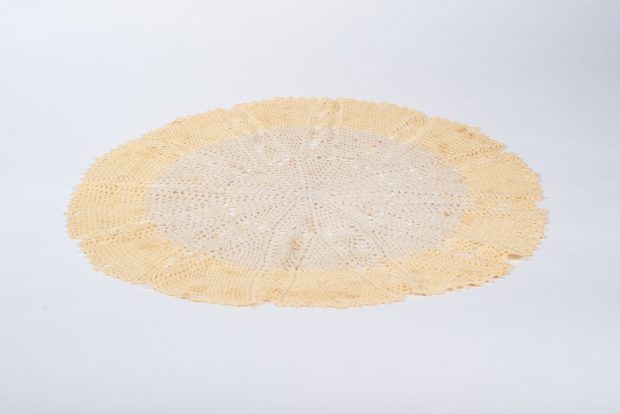 Centre de table circulaire crocheté déposé sur un fond blanc. Le tour est d’un jaune très pâle tandis que l’intérieur est écru. Les points forment un motif d’étoile à douze pointes.