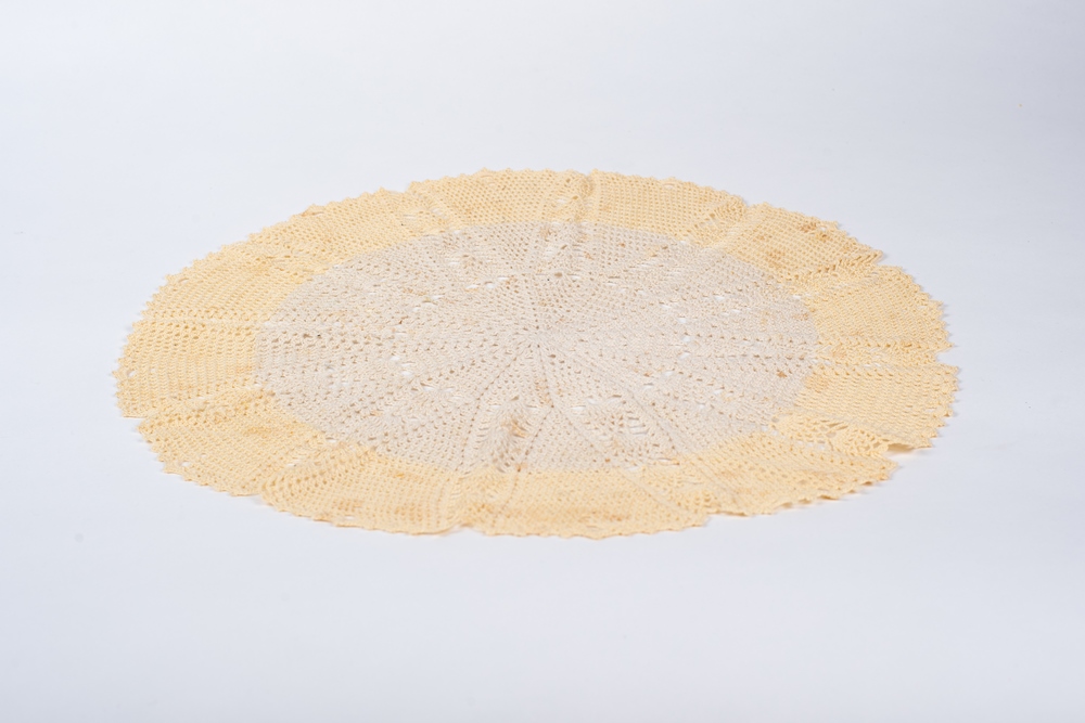 Centre de table circulaire crocheté déposé sur un fond blanc. Le tour est d’un jaune très pâle tandis que l’intérieur est écru. Les points forment un motif d’étoile à douze pointes.