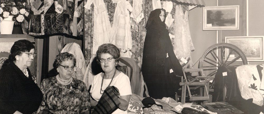 Photographie en noir et blanc de trois femmes examinant une couverture. Derrière elles, une courtepointe et quelques vêtements sont accrochés. À leur droite, des couvertures sont posées sur une table et un rouet est exposé.