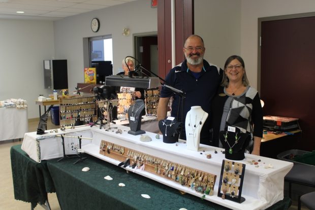 Un homme et une femme souriants sont debout derrière une table sur laquelle sont exposés des boucles d’oreilles et des colliers.