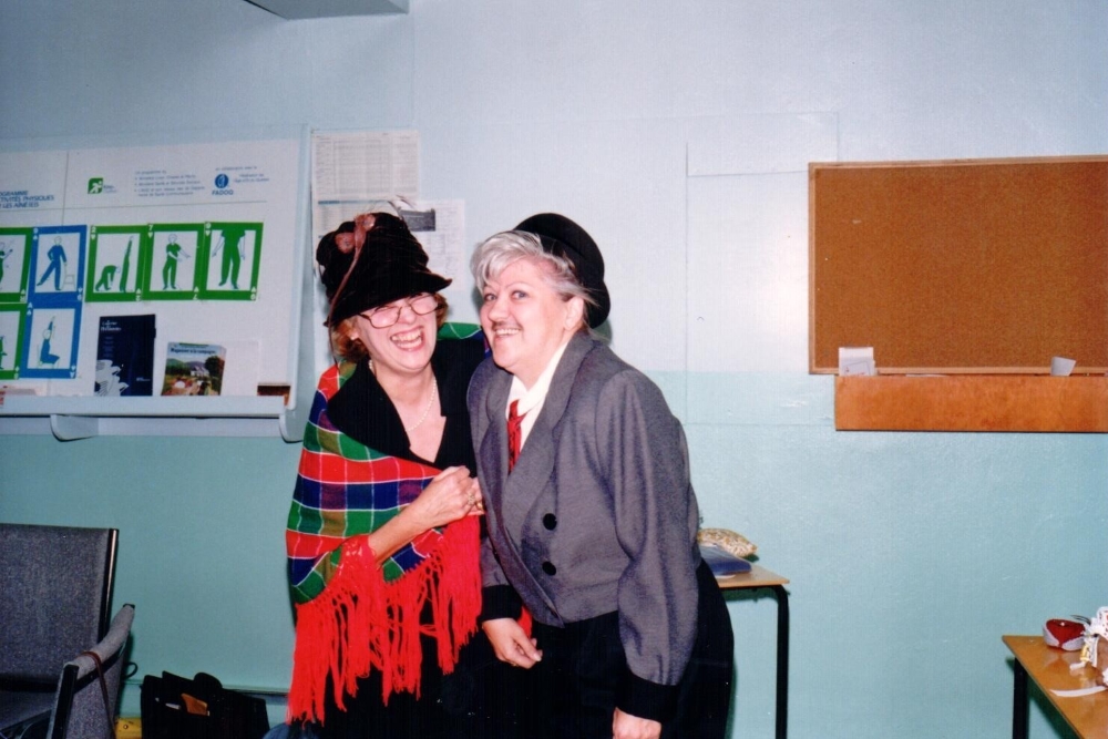 Deux femmes déguisées sont bras dessus bras dessous et rigolent. Celle de gauche porte un chapeau noir et un châle rouge, bleu et vert orné de franges rouges. Celle de droite porte un chapeau, un veston gris, une cravate et une fausse moustache.