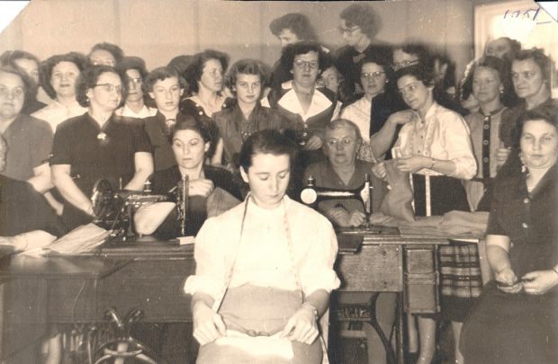 Photographie en noir et blanc. À l’avant-plan est assise une femme vêtue d’une chemise blanche et portant un ruban à mesurer autour de son cou. Derrière elle, on voit deux femmes installées derrière des machines à coudre. À l’arrière-plan, une vingtaine de femmes sont debout.  