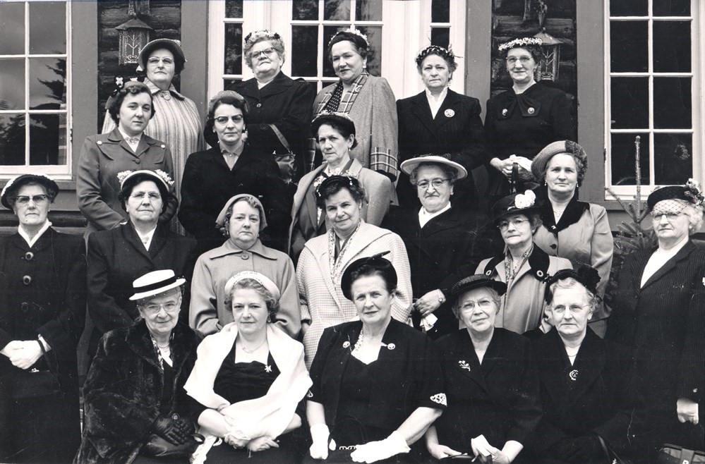 Photographie en noir et blanc prise devant un bâtiment. Vingt-et-une femmes portant chacune un veston ou un manteau et un chapeau forment quatre rangées. Les femmes de la première rangée sont assises tandis que les autres sont debout. 