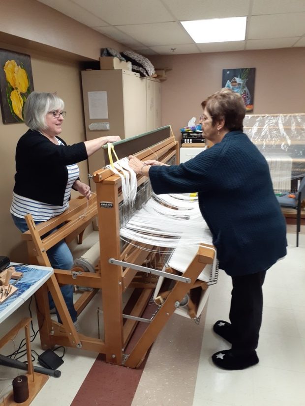 Deux femmes sont debout chacune d’un côté d’un métier à tisser de bois. Elles procèdent au montage du métier avec des fils blancs. À l’arrière-plan, on aperçoit un autre métier à tisser. 