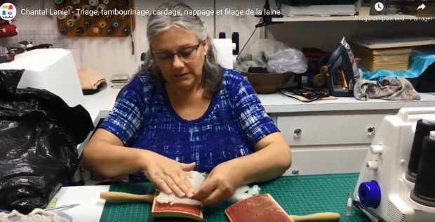 Une femme aux cheveux gris vêtue d'un chandail bleu est assise derrière une table sur laquelle est posée un tapis de coupe. Devant elle, se trouvent deux peignes à carder. À l'aide de ses mains, elle place de la laine blanche sur un des deux.