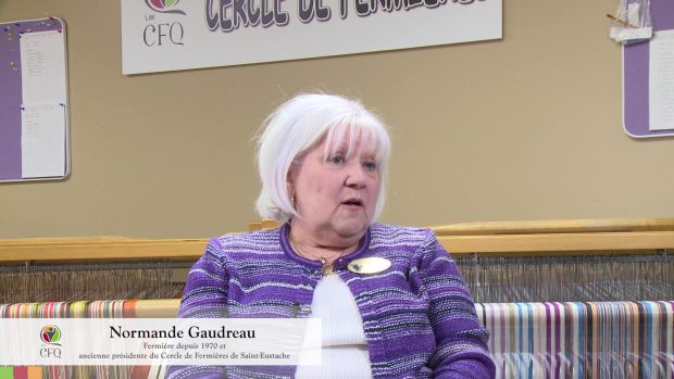 Une femme aux cheveux blancs vêtue d'un cardigan rayé violet est assise devant un métier à tisser. 