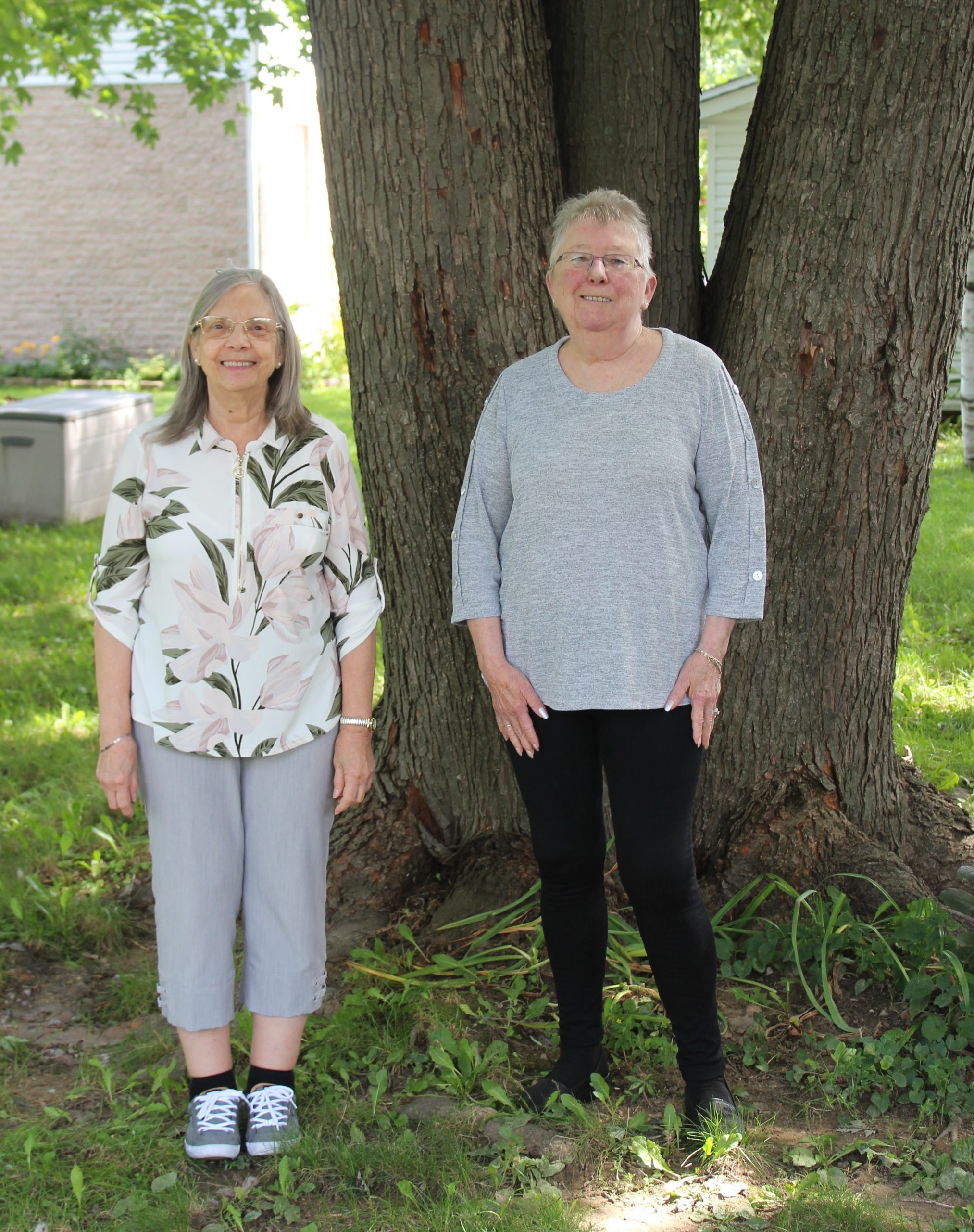 Deux femmes sont debout dehors devant un grand arbre en été. Elles regardent devant elles et sont souriantes.