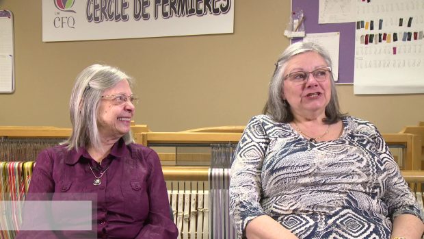 Deux femmes aux cheveux gris sont assises, côte à côte, devant un métier à tisser. Celle de gauche regarde celle de droite en souriant. Celle de droite parle.