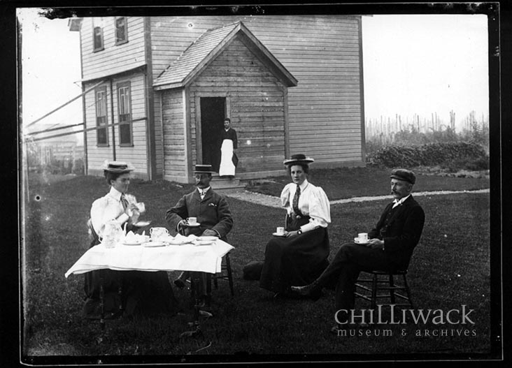 Portrait de deux femmes et deux hommes assis en train de prendre le thé devant la maison Hulbert avec un domestique chinois à l’arrière-plan 