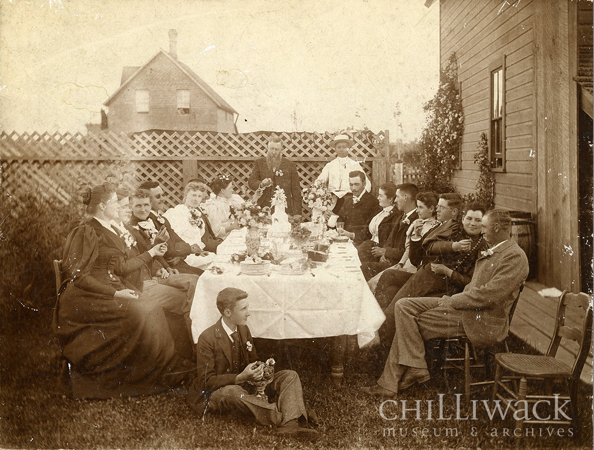 Photographie représentant un groupe de dix-huit personnes autour d’une table et une personne assise par terre. Maison et barrière en arrière-plan 