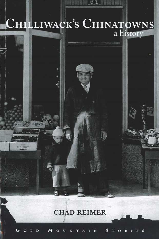 Couverture du livre du Dr Chad Reimer « Chilliwack’s Chinatowns: A History ». La photo de couverture en noir et blanc montre un jeune garçon et un homme se tenant la main, debout devant un magasin. On aperçoit un étal de fruits sur la gauche et un étal de légumes sur la droite.