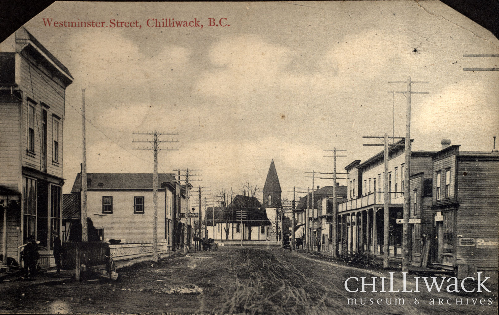Carte postale en noir et blanc de la rue Westminster, à Chilliwack, en Colombie-Britannique. La rue est composée d’édifices à étage avec une église en arrière-plan