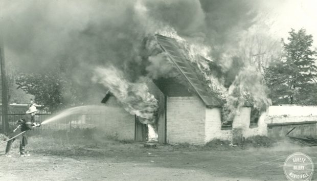 Image blanche et grise d'un feu engloutissant un petit bâtiment, il y a aussi un pompier pulvérisant de l'eau sur le bâtiment.