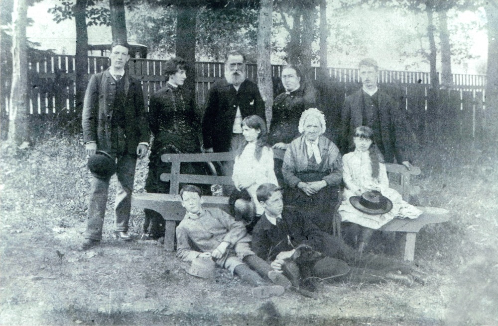 Photo noir et blanc d'une famille dans une cour entourée d'arbres et d'une clôture en bois. Dans cette image, il y a dix personnes. Cinq personnes sont debout, trois sont assis sur un banc, et deux sont couchés sur le sol dans l'herbe. Ils s'habillent tous leurs meilleurs dimanche.