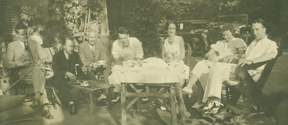 image sépia de gens assis sur des chaises autour d'un ensemble de thé sur une table recouverte de lin blanc
