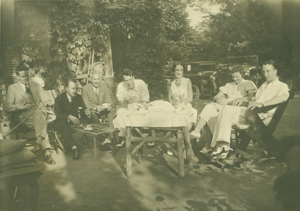 image sépia de gens assis sur des chaises autour d'un ensemble de thé sur une table recouverte de lin blanc