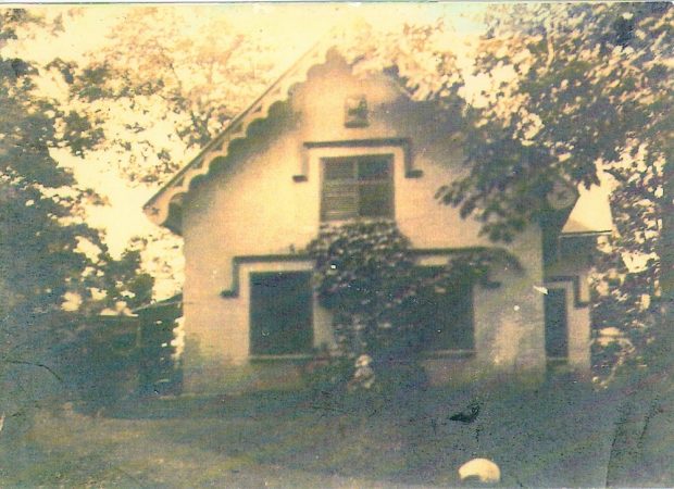 Image grise et blanche d'une maison blanche avec des détails coloniaux anglais entourée d'arbres et de buissons.