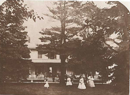 Photo jaune et orange d'une maison par enveloppées arbres conifères. Il y a des femmes vêtues de robes blanches devant la maison sur l'herbe par un grand arbre.