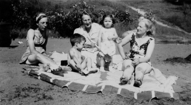 photo en noir et blanc d'un groupe de gens assis sur la plage. Il y a trois femmes vêtues de vêtements de plage et un petit garçon et une fille. Ils sont tous sur une grande feuille blanche.