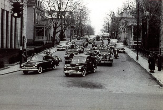 photo en noir et blanc d'un cortège funèbre, de nombreuses voitures noires conduisez sur une route.