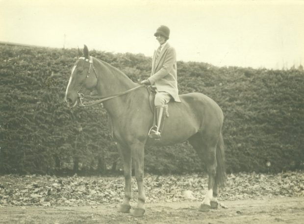 photo sépia d'une femme assise sur un grand cheval, ils sont debout devant une haie