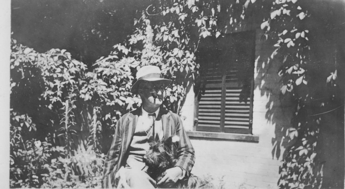 Photo noir et blanc d'un vieil homme vêtu d'un costume ample avec une pipe à la bouche et un chien Scottie noir sur ses genoux. Ils sont assis sur une chaise devant la maison et sont entourés de vignes.