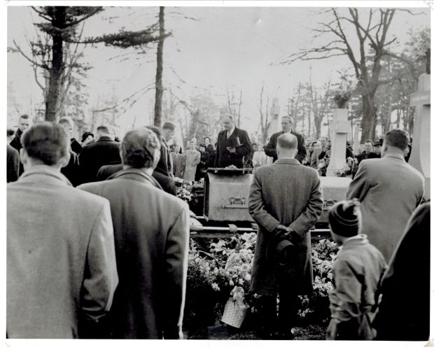 photo en noir et blanc d'un service funéraire. Les hommes sont debout avec leur tête baissée autour d'un cercueil descendu dans le sol avec un petit garçon curieux en arrière-plan. Fleurs et pierre tombale de granit entourent le cercueil.