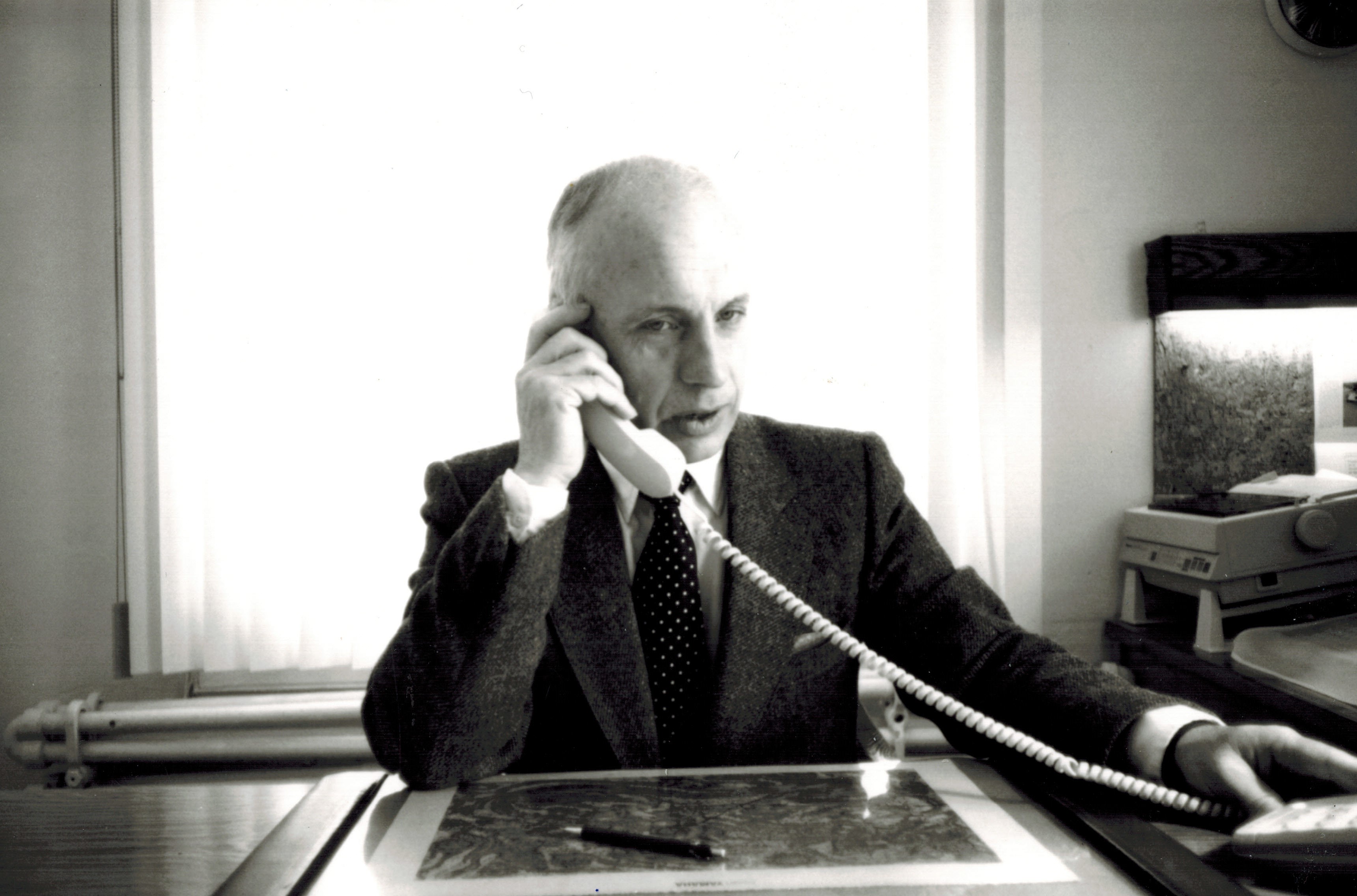 photo en noir et blanc d'un homme vêtu d'un costume tenant un téléphone à son oreille dans un bureau