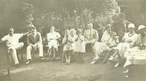 Une image sépia des hommes, des femmes et des enfants assis en demi-cercle qui portent des vêtements somptueux à une partie de thé de famille Treggett entouré d'un paysage verdoyant de vignes vertes
