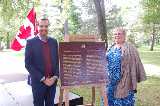Homme et femme debout de part et d'autre d'une plaque commémorative du lieu historique national du Canada avec un drapeau canadien à l'arrière-plan