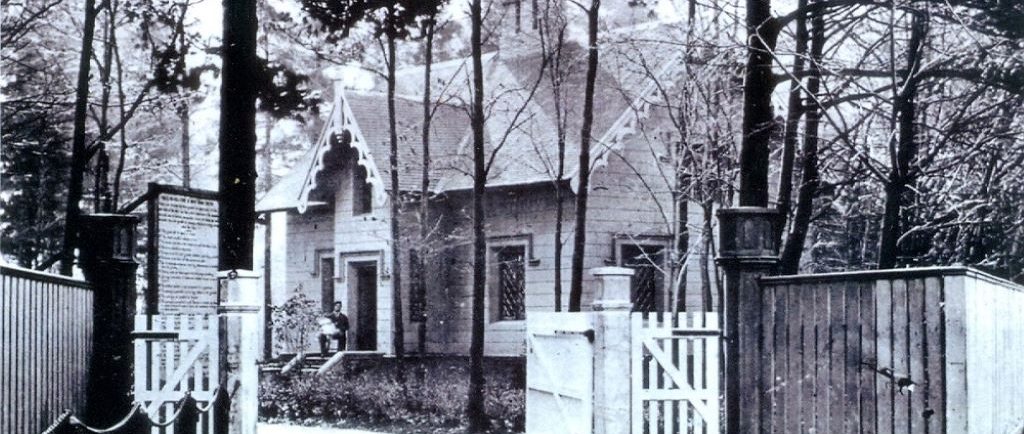 photo en noir et blanc d'une maison dans les bois. Un homme est assis sur l'étape avant la tenue d'un bébé vêtu blanc. Il y a aussi une grande clôture de bois devant la maison.