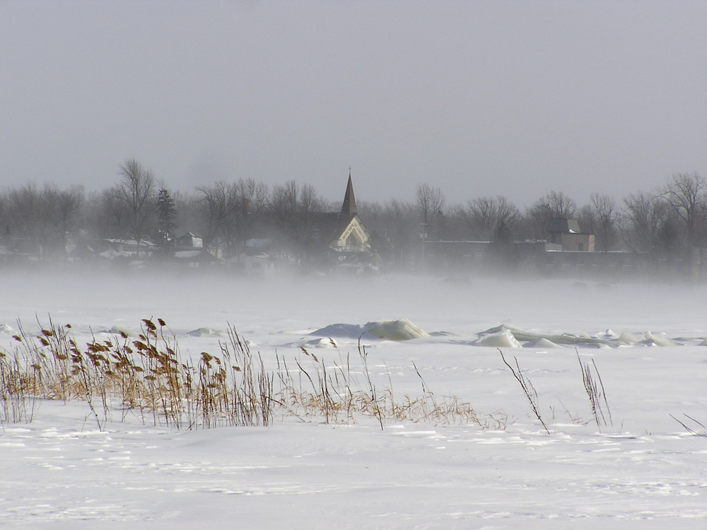 Photographie couleur prise en hiver, plan éloigné, vue d’un village au bord d’une rivière gelée, au centre, le clocher d’une église visible dans la poudrerie.