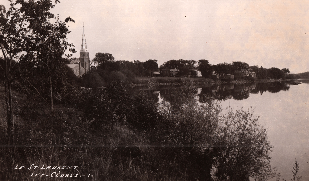 Photographie ancienne en noir et blanc, plan éloigné, en avant-plan, une vaste étendue d’eau, des arbres et de la végétation, en arrière-plan, le clocher d’une église avec à sa droite une série de petites maisons. 