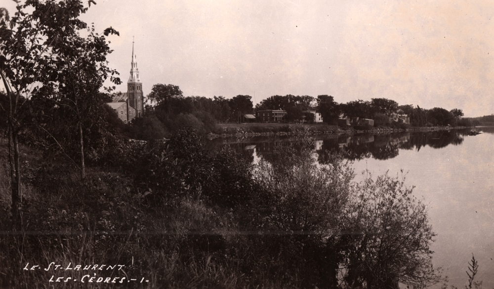 Photographie ancienne en noir et blanc, plan éloigné, en avant-plan, une vaste étendue d’eau, des arbres et de la végétation, en arrière-plan, le clocher d’une église avec à sa droite une série de petites maisons.