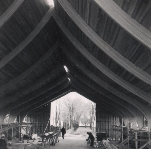 Photographie en noir et blanc, plan rapproché d’une charpente en bois d’un bâtiment en construction dont l’arrière est ouvert, à l’intérieur des matériaux de construction et des ouvriers, en avant-plan, à la gauche, une femme regarde la caméra.