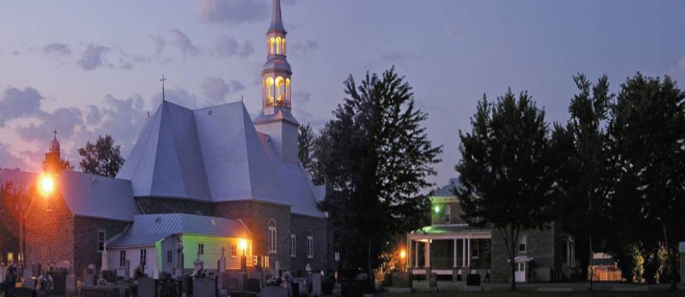Photographie couleur prise de nuit, plan éloigné de l’arrière des bâtiments comprenant l’église, le presbytère et le cimetière, l’église est éclairée.